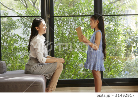 A little girl is holding a book and standing in front of her mother. 31492917