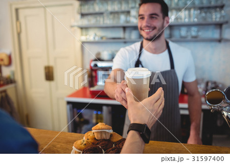 Happy barista offering coffee in disposable cup to customer at cafe Happy barista offering coffee in disposable cup to customer at cafe 31597400