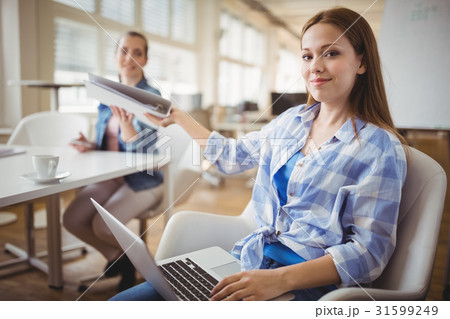Businesswoman giving file to colleague while working on laptop at office Businesswoman giving file to colleague while working on laptop at office 31599249