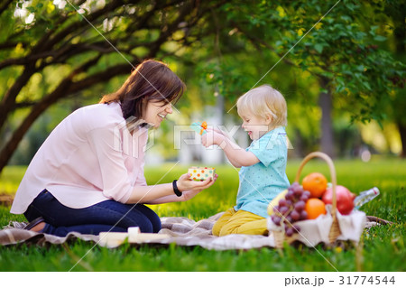 Cute little boy with his young mother opening nicely wrapped gift during picnic in sunny park 31774544