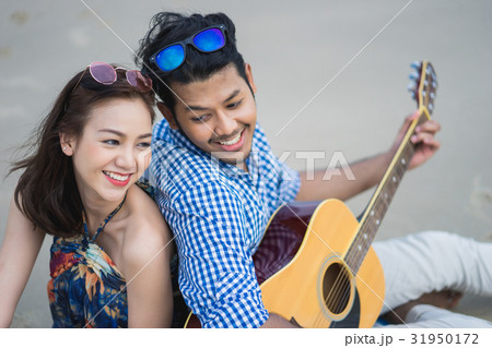 A picture of a happy couple sitting and playing guitar on the beach 31950172
