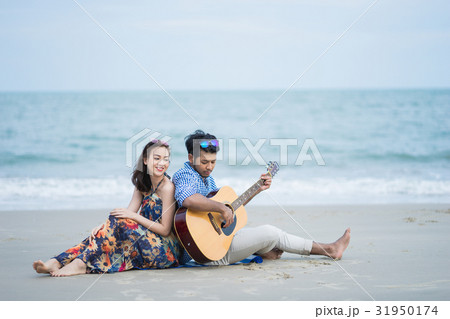 A man is sitting close his girlfriend and playing guitar on the beach A man is sitting close his girlfriend and playing guitar on the beach 31950174