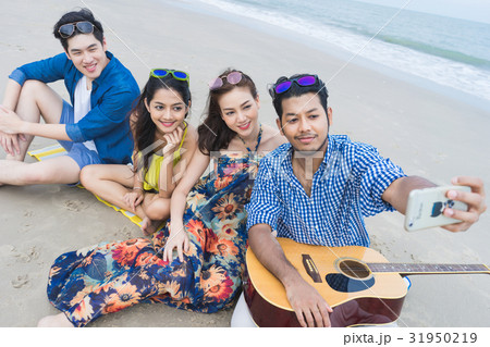 four people are sitting, smiling and taking selfie on the beach 31950219