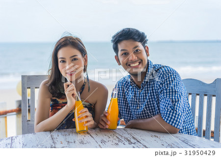A photo of a couple sitting, smiling, holding beverages near a beach. 31950429