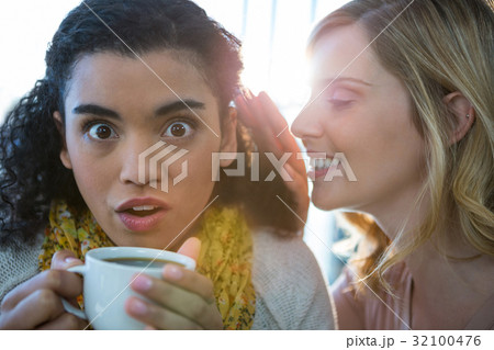 Woman whispering a secret into her friends ear while having coffee Woman whispering a secret into her friends ear while having coffee 32100476