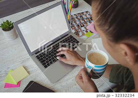 Female photo editor holding coffee cup while using laptop in office Female photo editor holding coffee cup while using laptop in office 32332234