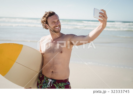 Smiling shirtless man taking selfie with surfboard at beach 32333745