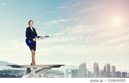Attractive businesswoman on metal tray playing electric guitar against cityscape background 33064095