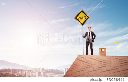 Young engineer on house brick roof holding yellow signboard and looking at city. Mixed media Young engineer on house brick roof holding yellow signboard and looking at city. Mixed media 33068441