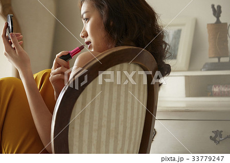 An Asian young woman is sitting on the chair and using the lipstick. 33779247