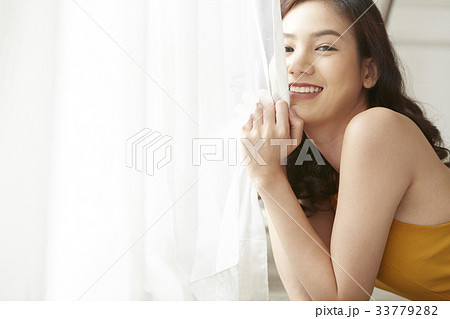 An Asian young woman is holding the curtain while smiling inside. 33779282
