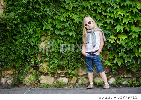 Funny little girl wearing sunglasses posing by bindweed wall on warm and sunny summer day 33779715
