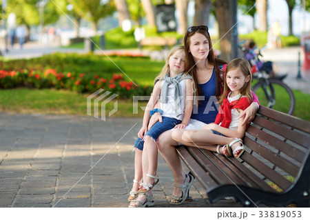 Young mother and two cute little sisters sitting on a bench hugging on warm and sunny summer day Young mother and two cute little sisters sitting on a bench hugging on warm and sunny summer day 33819053