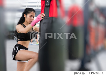 young woman execute exercise in fitness center. female athlete hitting and punching sandbag with rowing machine in boxing gym. sporty girl working out in health club. martial arts, muay thai concept young woman execute exercise in fitness center. female athlete hitting and punching sandbag with rowing machine in boxing gym. sporty girl working out in health club. martial arts, muay thai concept 33928684