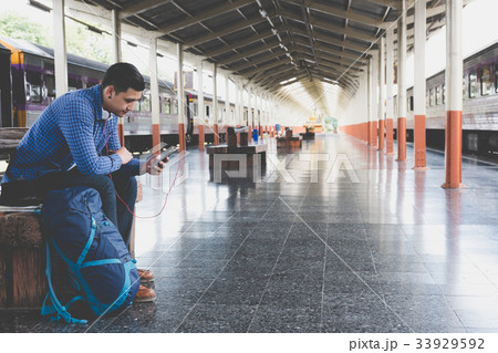 asian man with backpack sitting on platform at train station. backpacker or traveler with headphone look at mobile phone while waiting for train. journey, trip, travel concept 33929592