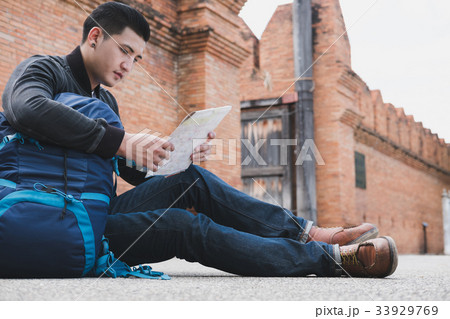 young traveler reading map. asian man wearing black jacket and blue jeans sitting near old orange brick wall with backpack 33929769