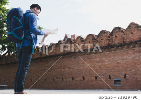 young traveler reading map. asian man wearing blue shirt and jeans standing near old orange brick wall with backpack 33929790
