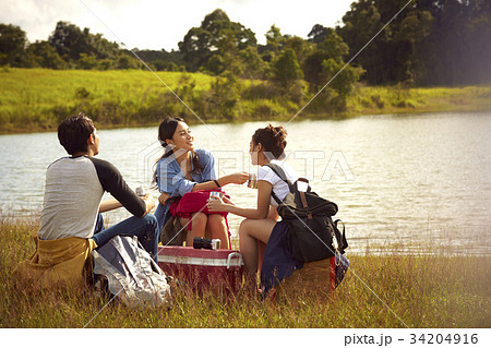 Three friends are sitting and drinking coffee on the riverbank. 34204916