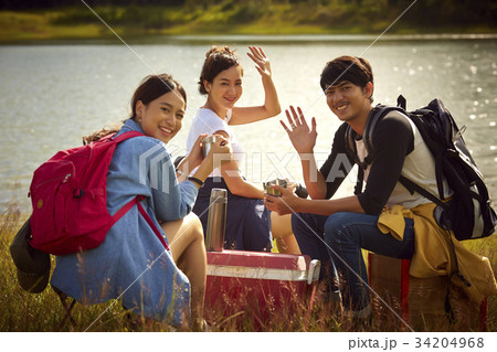 A group of friends is sitting, smiling and waving hand on the riverside 34204968