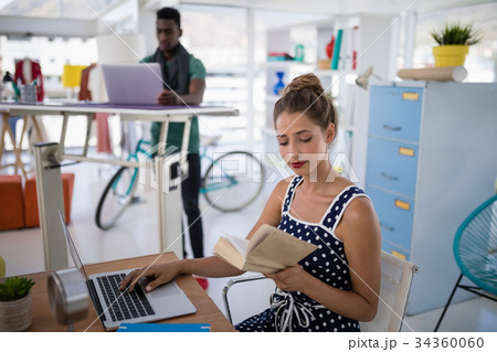 Female executive working on laptop while reading a book at desk Female executive working on laptop while reading a book at desk 34360060