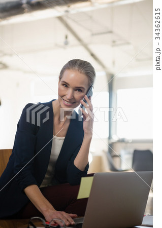 Portrait of woman talking on phone while using laptop in office 34362615
