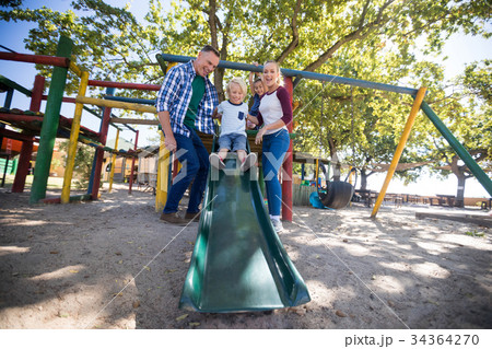 Happy parents standing by children playing on slide at playground Happy parents standing by children playing on slide at playground 34364270