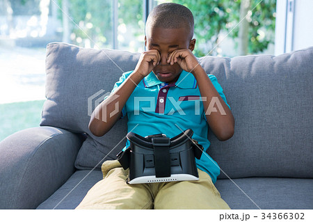 Boy rubbing eyes while sitting with VR headset on sofa Boy rubbing eyes while sitting with VR headset on sofa 34366302