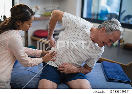Female therapist examining back of senior male patient sitting on bed Female therapist examining back of senior male patient sitting on bed 34366947