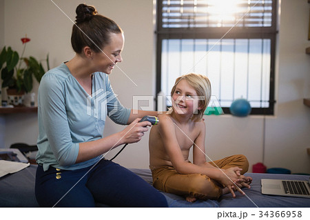 Smiling young female therapist scanning shoulder while looking at shirtless boy sitting on bed Smiling young female therapist scanning shoulder while looking at shirtless boy sitting on bed 34366958