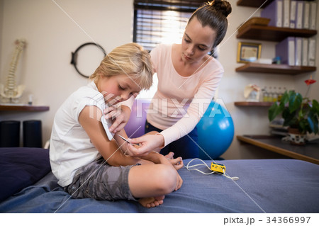 Young female therapist positioning electrodes on arm of boy Young female therapist positioning electrodes on arm of boy 34366997