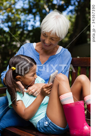 Cheerful senior woman enjoying with girl holding mobile phone on bench 34367390