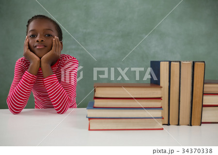Schoolgirl sitting beside books stack against chalkboard 34370338