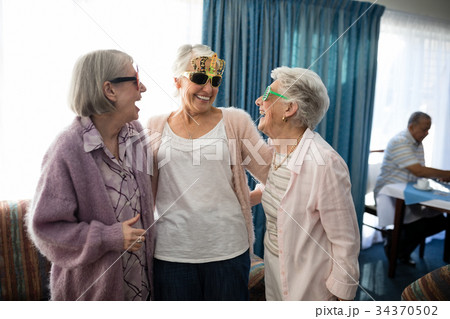Senior female friends wearing novelty glasses talking at nursing home Senior female friends wearing novelty glasses talking at nursing home 34370502