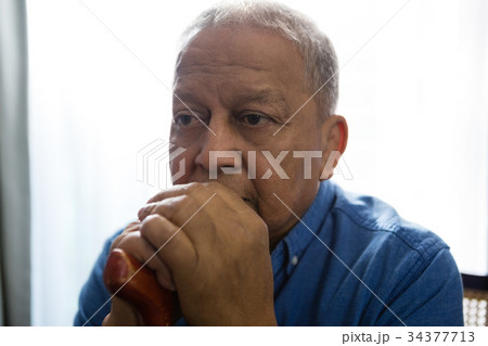 Thoughtful sad senior man holding walking cane while sitting on chair Thoughtful sad senior man holding walking cane while sitting on chair 34377713