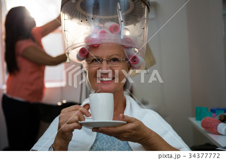 Portrait of senior woman having drink while sitting under hair steamer 34377772