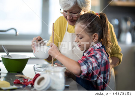 Grandmother adding water while granddaughter is mixing flour in a bowl 34378205