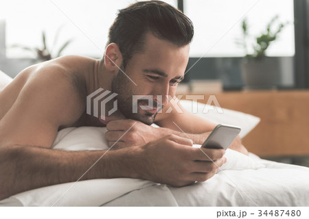 Joyful guy messaging on mobile phone in bedroom Joyful guy messaging on mobile phone in bedroom 34487480