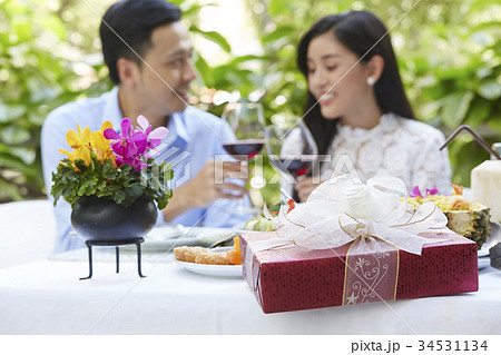 Two people are sitting on a table with food and gift and holding glasses of wine 34531134
