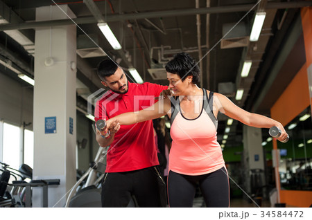 Fitness coach assisting his female client in gym Fitness coach assisting his female client in gym 34584472