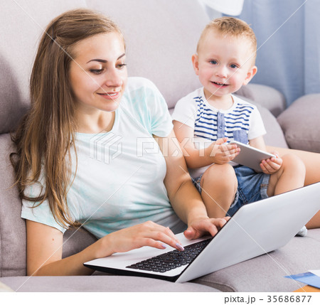 Smiling mother is productively working behind laptop while child playing on tablet Smiling mother is productively working behind laptop while child playing on tablet 35686877