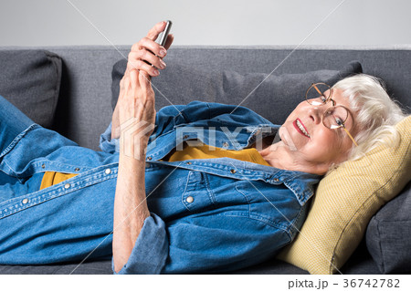 Aged cheerful lady lying on sofa with mobile phone Aged cheerful lady lying on sofa with mobile phone 36742782