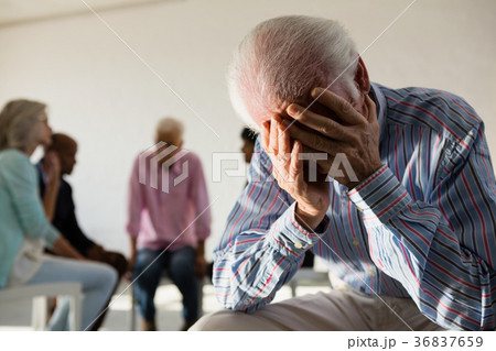 Senior man with head in hand sitting on hair with friends discussing in background Senior man with head in hand sitting on hair with friends discussing in background 36837659