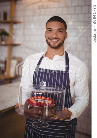 Portrait of smiling young waiter holding cake on glass cakestand 36841731