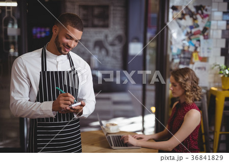 Smiling young waiter writing in notepad while standing at coffee shop Smiling young waiter writing in notepad while standing at coffee shop 36841829
