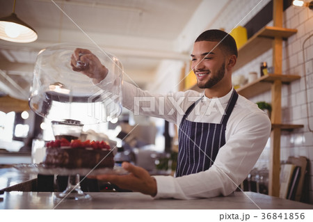 Smiling young waiter holding glass lid over cakestand at counter 36841856