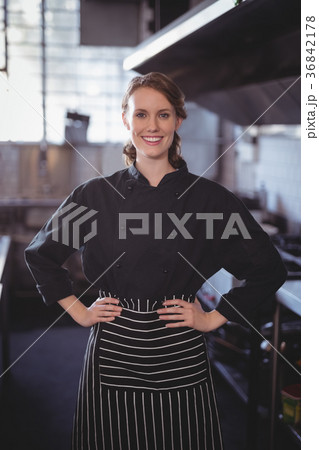 Portrait of smiling young female barista standing with hands on hip in kitchen Portrait of smiling young female barista standing with hands on hip in kitchen 36842178