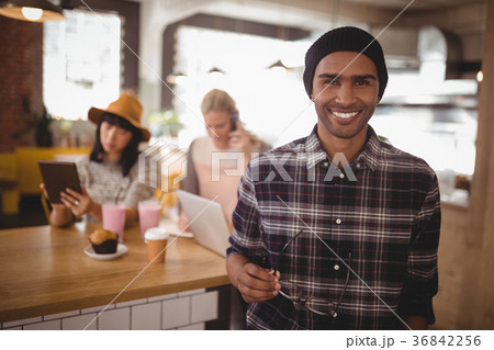 Portrait of smiling young man holding eyeglasses standing against female friends at coffee shop 36842256