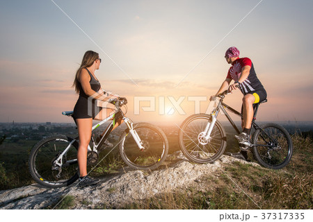 Cyclist couple with mountain bikes on the hill at sunset Cyclist couple with mountain bikes on the hill at sunset 37317335