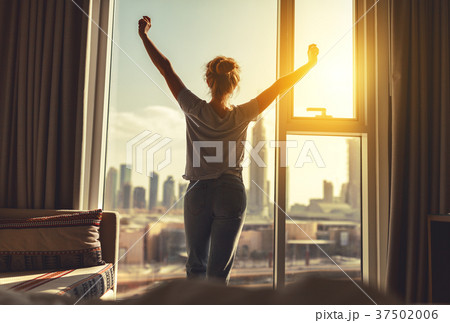 happy woman stretches and  opens curtains at window in morning 37502006