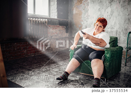 Overweight woman sits in a chair and watches TV 37555534